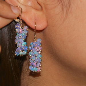 May include: A pair of long, dangling earrings with a cluster of iridescent opal beads in shades of blue, purple, and pink. The beads are strung on a gold chain and the earrings have a simple gold hook closure.