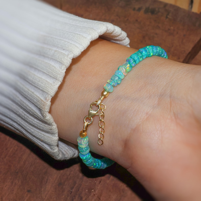 May include: A delicate bracelet made with light blue opal beads and a gold clasp. The bracelet is shown on a person's wrist.