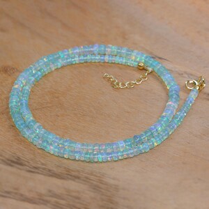 May include: A delicate necklace made of small, iridescent opal beads. The beads are a pale blue colour with flashes of rainbow hues. The necklace has a gold-coloured chain and clasp. The necklace is displayed on a wooden surface.