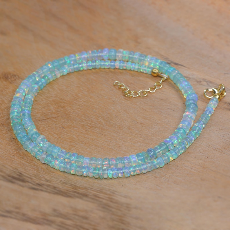 May include: A delicate necklace made of small, iridescent opal beads. The beads are a pale blue colour with flashes of rainbow hues. The necklace has a gold-coloured chain and clasp. The necklace is displayed on a wooden surface.