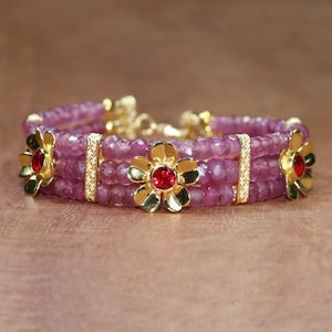 May include: A multi-strand bracelet featuring faceted pink beads arranged in three rows. The design includes three gold-toned metal flower charms, each centred with a small, round, vibrant red stone. Gold-toned textured spacer bars separate the rows.