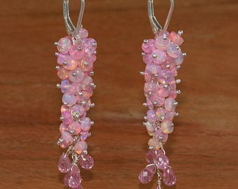 May include: Dangle earrings with pink opal beads and teardrop-shaped pink gemstones. The earrings have silver-coloured leverback closures. The design is set against a wooden background.