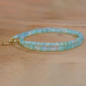May include: A delicate necklace featuring small, iridescent opal beads. The necklace is a soft blue-green colour and has a gold-coloured clasp. The beads are strung together to create a layered effect, resting on a light brown wooden surface.