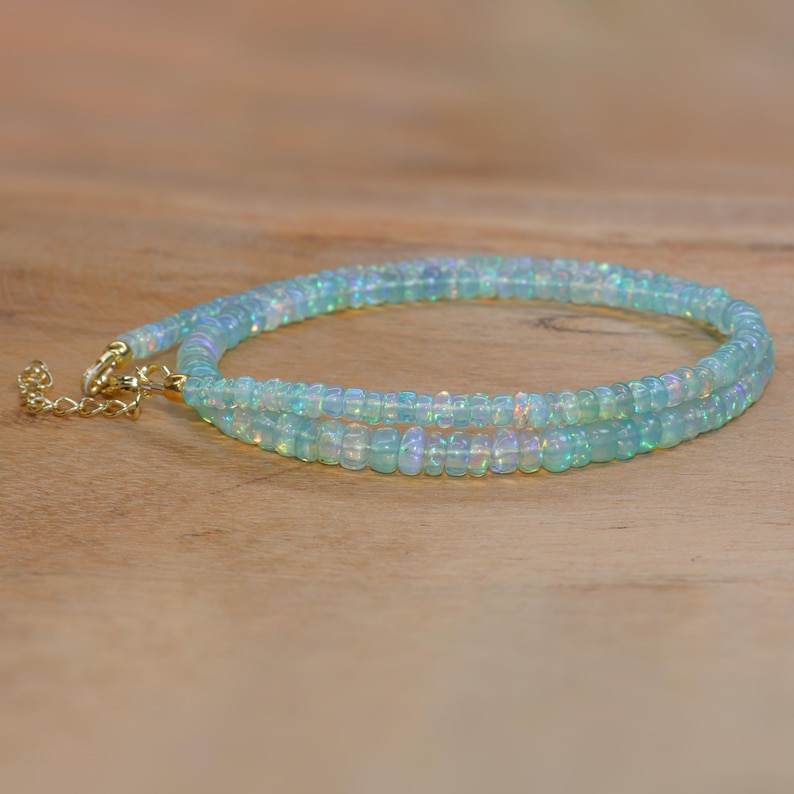 May include: A delicate necklace featuring small, iridescent opal beads. The necklace is a soft blue-green colour and has a gold-coloured clasp. The beads are strung together to create a layered effect, resting on a light brown wooden surface.
