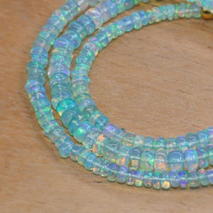 May include: Close-up of three strands of iridescent opal beads. The beads are a light blue colour with flashes of rainbow hues. The beads are strung together, creating a necklace or bracelet.