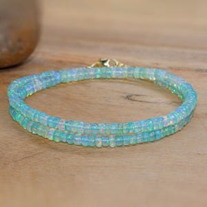 May include: A delicate, double-strand bracelet composed of small, iridescent, light blue beads. The bracelet is secured with a gold-colored clasp. The beads have a subtle, shimmering quality, and the bracelet is displayed on a wooden surface.