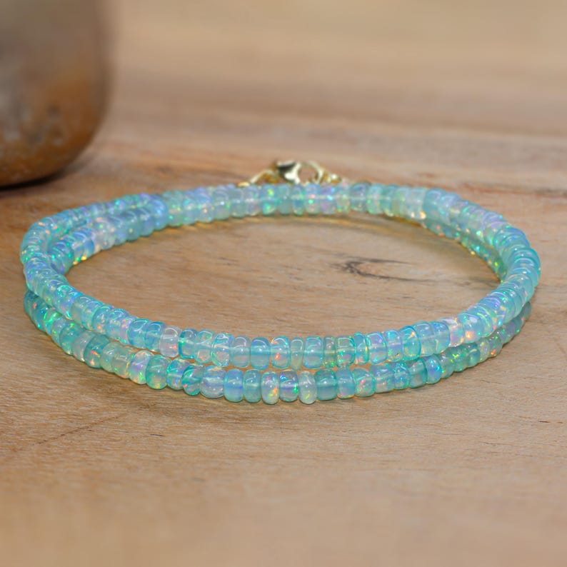 May include: A delicate, double-strand bracelet made of small, iridescent, light blue beads. The bracelet is secured with a gold-coloured clasp. The beads have a subtle, shimmering quality, and the bracelet is displayed on a wooden surface.