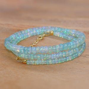 May include: A delicate necklace featuring iridescent, light blue opal beads. The necklace is strung on a gold-coloured chain with a lobster clasp and an extender chain. The beads are small and round, creating a layered effect. The necklace is displayed on a wooden surface.