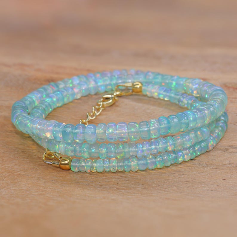 May include: A delicate necklace featuring iridescent, light blue opal beads. The necklace is strung on a gold-coloured chain with a lobster clasp and an extender chain. The beads are small and round, creating a layered effect. The necklace is displayed on a wooden surface.