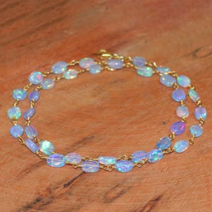 May include: A layered necklace with oval opals in blue, pink, and white. The opals are strung on a gold-coloured chain. The necklace is displayed on a wooden surface. The design of the necklace is highlighted by the wooden background.
