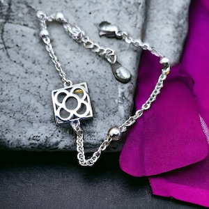 Può includere: Un braccialetto d'argento con un ciondolo quadrato che presenta un delicato design floreale. Il ciondolo è realizzato in metallo traforato e presenta un design delicato e intricato.