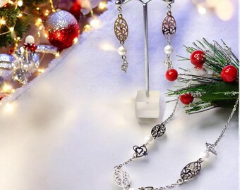 May include: A silver necklace and earrings set with delicate filigree designs and white pearls. The necklace features a chain with oval and heart-shaped pendants. The earrings are drop earrings with oval pendants.