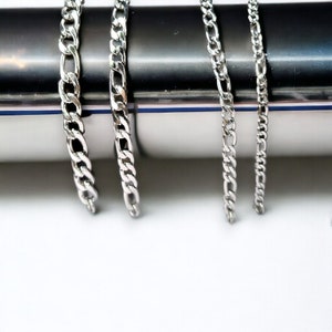 Könnte beinhalten: Vier silberne Ketten in verschiedenen Breiten und Längen, die vor einem silbernen Hintergrund hängen.