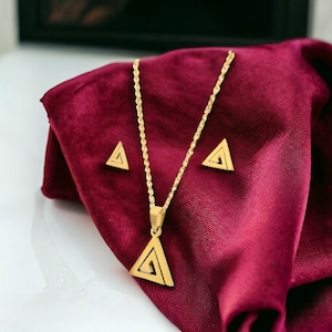 May include: A gold necklace with a triangle pendant and matching triangle earrings. The necklace is on a red velvet background.