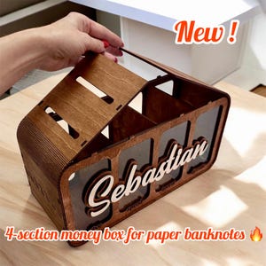 May include: A wooden money box with four sections labeled "Save", "Spend", "Invest", and "Donate". The box has a clear plastic front and a hinged lid. The box is personalized with the name "Sebastian".