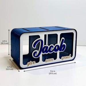 Peut inclure: Tirelire en bois bleu avec trois compartiments &eacute;tiquet&eacute;s "give", "save" et "spend". La tirelire est personnalis&eacute;e avec le nom "Jacob" en lettres blanches et bleues.