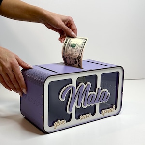 Peut inclure: Une bo&icirc;te &agrave; monnaie en bois violette avec trois fentes &eacute;tiquet&eacute;es "donner", "&eacute;pargner" et "d&eacute;penser". La bo&icirc;te a un devant en plastique transparent et le nom "Maia" est &eacute;crit en blanc sur le devant.