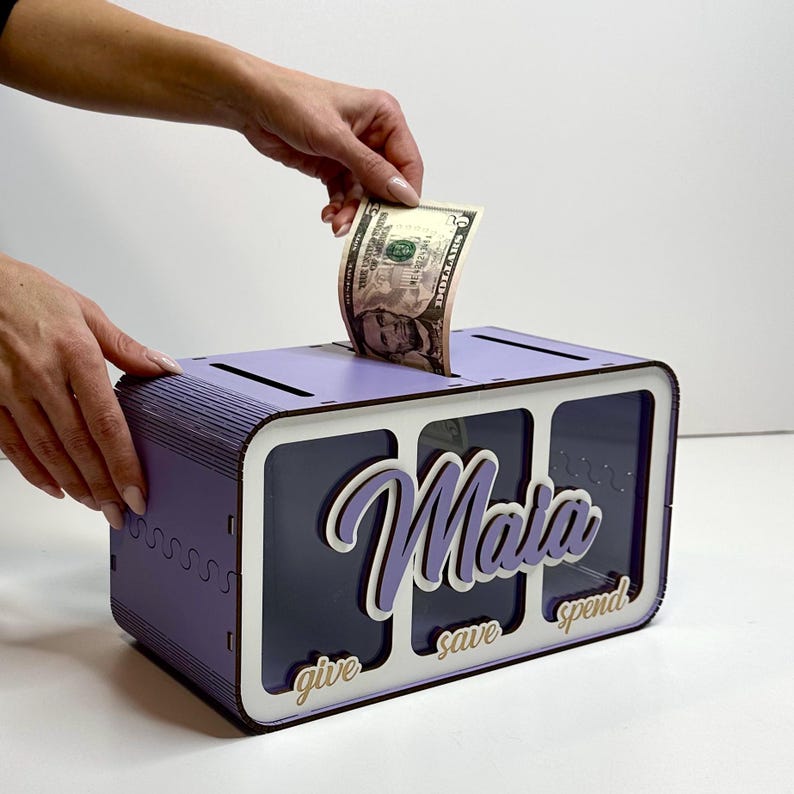 Peut inclure: Une bo&icirc;te &agrave; monnaie en bois violette avec trois fentes &eacute;tiquet&eacute;es "donner", "&eacute;pargner" et "d&eacute;penser". La bo&icirc;te a un devant en plastique transparent et le nom "Maia" est &eacute;crit en blanc sur le devant.