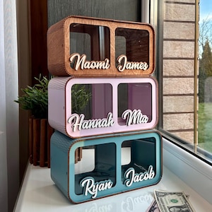 Puede incluir: Un conjunto de tres cajas de madera para guardar dinero apiladas una encima de la otra. La caja superior es marrón, la caja del medio es rosa y la caja inferior es azul. Cada caja tiene dos compartimentos con un nombre en cada compartimento. Los nombres en las cajas son Naomi, James, Hannah, Mia, Ryan y Jacob.