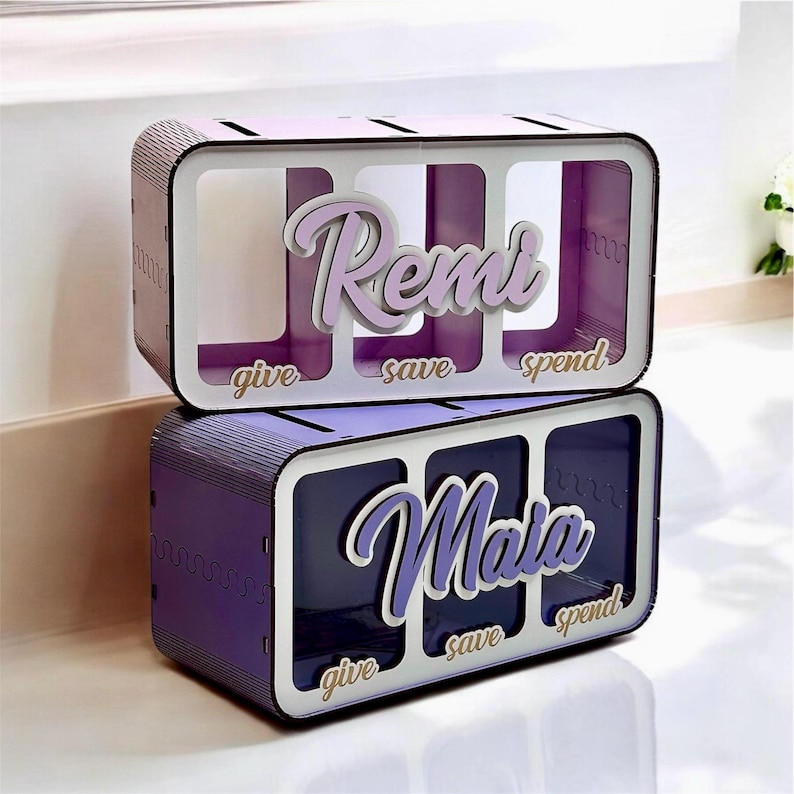 Peut inclure: Deux bo&icirc;tes &agrave; monnaie en bois avec trois compartiments chacune. La bo&icirc;te du haut est violette clair avec le nom "Remi" et les mots "donner", "&eacute;pargner" et "d&eacute;penser". La bo&icirc;te du bas est violette fonc&eacute; avec le nom "Maia" et les mots "donner", "&eacute;pargner" et "d&eacute;penser".