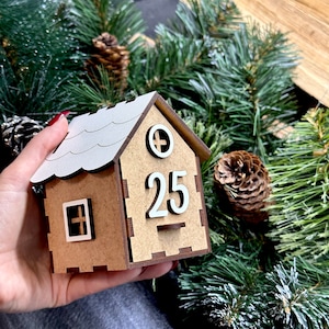 May include: A wooden house-shaped advent calendar with a white roof and a white number 25 on the front. The house is brown and has a small window with a white cross.