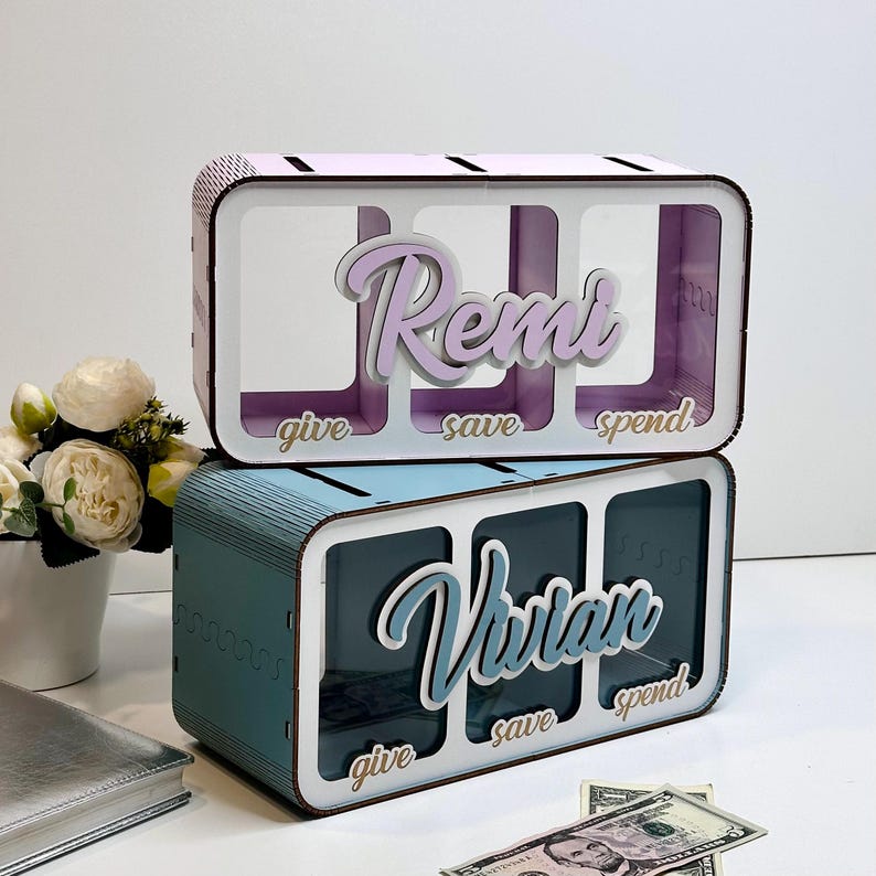 Peut inclure: Deux tirelires en bois avec trois fentes chacune. La tirelire du haut est blanche avec un nom rose "Remi" et les fentes sont &eacute;tiquet&eacute;es "donner", "&eacute;pargner" et "d&eacute;penser". La tirelire du bas est bleue avec un nom bleu "Vivian" et les fentes sont &eacute;tiquet&eacute;es "donner", "&eacute;pargner" et "d&eacute;penser".