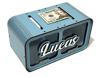 May include: A light blue wooden coin bank with two compartments labeled "save" and "spend". The bank has a slot on the top for inserting money and features the name "Lucas" in white lettering. A $20 bill is visible in the slot.