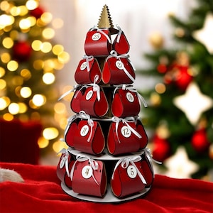 Può includere: Un calendario dell'Avvento rosso a forma di albero di Natale. Ogni sacchetto è numerato e legato con un nastro bianco. L'albero è su una base circolare bianca, sormontato da una cima dorata.