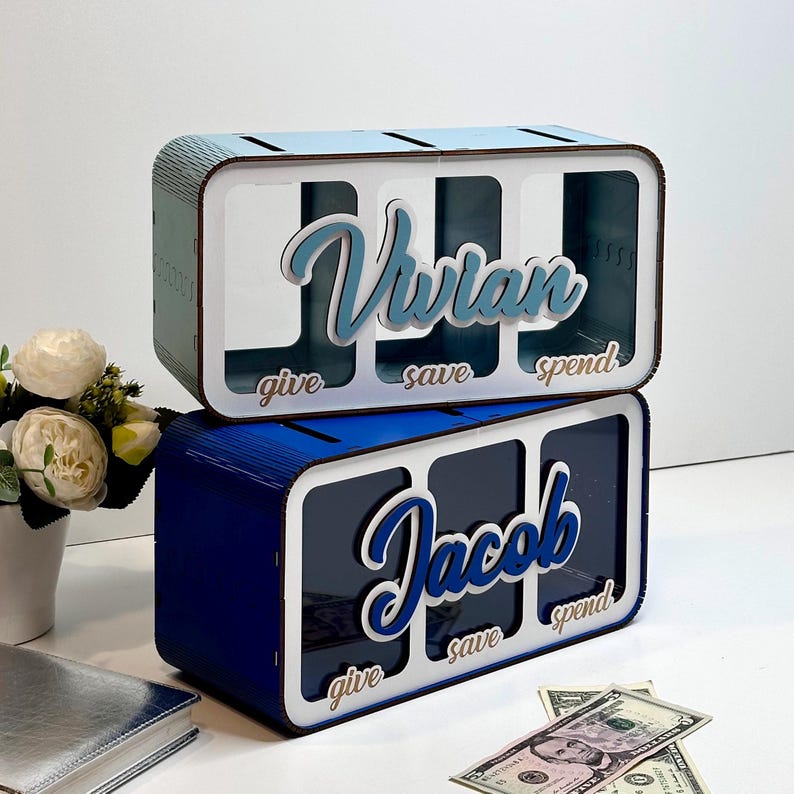 Peut inclure: Deux bo&icirc;tes &agrave; monnaie en bois peintes en bleu et blanc. La bo&icirc;te du haut est nomm&eacute;e "Vivian" et la bo&icirc;te du bas est nomm&eacute;e "Jacob". Chaque bo&icirc;te a trois fentes &eacute;tiquet&eacute;es "donner", "&eacute;pargner" et "d&eacute;penser".