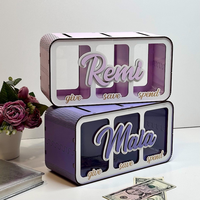 Peut inclure: Tirelire en bois violet avec trois fentes &eacute;tiquet&eacute;es "donner", "&eacute;pargner" et "d&eacute;penser". La partie sup&eacute;rieure porte le nom "Remi" et la partie inf&eacute;rieure porte le nom "Maia".