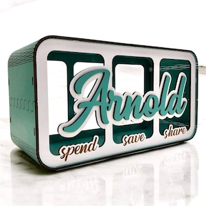 Puede incluir: Un organizador de dinero de madera en color verde azulado y blanco con el nombre "Arnold" en escritura turquesa. El organizador tiene tres compartimentos etiquetados como "spend", "save" y "share". Los bordes están delineados en marrón.