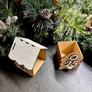 May include: A wooden Christmas advent calendar shaped like a house with a white roof. The calendar has a small door with the number 25 on it.