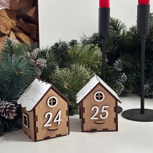 May include: Two wooden Christmas countdown houses with white roofs and numbers 24 and 25 on the front. The houses are sitting on a table with a green pine branch in the background.