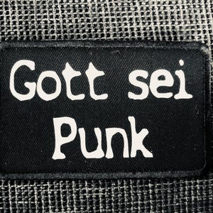 Könnte beinhalten: Ein rechteckiger schwarzer Aufnäher mit gesticktem Rand, der den weißen Schriftzug Gott sei Punk in einer stilisierten Schriftart zeigt. Der Aufnäher hat eine flache, stoffartige Textur und ist vor einem dunklen, gewebten Gitterhintergrund zentriert.