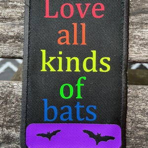 Könnte beinhalten: Ein rechteckiger, schwarz bestickter Aufnäher mit abgerundeten Ecken, der den Text Love in Rot, all in Orange, kinds in Gelb, of in Grün und bats in Blau zeigt. Unter dem Text befindet sich ein violetter horizontaler Balken mit zwei schwarzen Fledermaussilhouetten.