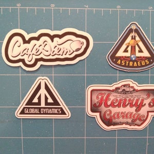 May include: Four stickers with different designs. The first sticker is white with black text that says "Cafe Diem" with a small coffee cup icon. The second sticker is a black triangle with a white rocket and planet inside. The text "ASTRAEUS" is at the bottom of the triangle. The third sticker is a black triangle with a white symbol inside. The text "GLOBAL DYNAMICS" is at the bottom of the triangle. The fourth sticker is a white rectangle with a red and white text that says "Henry's Garage" with a vintage truck graphic.