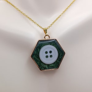 Puede incluir: Un collar de cadena dorada con un colgante hexagonal que presenta un botón blanco con cuatro agujeros sobre un fondo verde marmoleado.