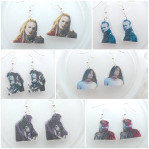 May include: A set of 10 pairs of dangle earrings featuring various images of musicians and actors. The earrings are made of clear plastic with silver hooks.