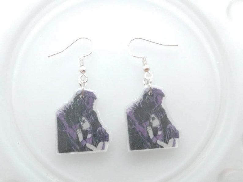 May include: A pair of silver dangle earrings with a purple and black image of a couple embracing. The image is printed on a clear plastic charm.