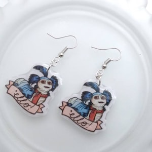 May include: A pair of dangle earrings featuring a cartoon character with blue hair, a red scarf, and the word "hello!" on a banner. The earrings have silver-colored hooks and are set against a white background.