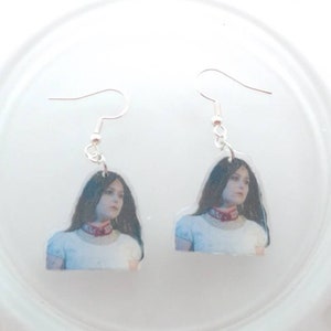 May include: A pair of silver dangle earrings with a photo of a woman with long dark hair wearing a white shirt and a red choker.