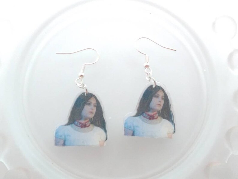 May include: A pair of silver dangle earrings with a photo of a woman with long dark hair wearing a white shirt and a red choker.