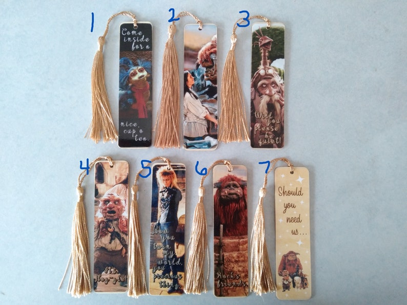 K&ouml;nnte beinhalten: Satz von sieben Lesezeichen mit Figuren und Zitaten aus dem Film Labyrinth. Jedes Lesezeichen hat einen goldenen Quasten. Die Lesezeichen zeigen Figuren wie Jareth, Sarah, Ludo und den Goblin-K&ouml;nig.