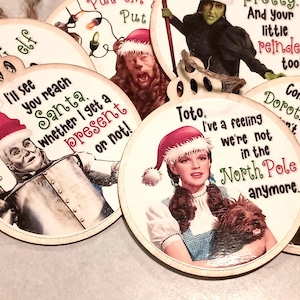 May include: Wooden Christmas ornaments featuring characters from The Wizard of Oz wearing Santa hats. Each ornament has a quote from the movie. The ornaments are round with a light wood grain finish and twine hangers.