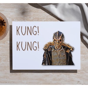 May include: A white greeting card with the words "KUNG! KUNG!" in brown letters. The card features an illustration of an alien-like figure wearing a jacket with a fur collar and a plaid shirt.