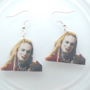 May include: A pair of dangle earrings featuring a printed image of a person with long blonde hair. The earrings are made of clear acrylic and have silver hooks.