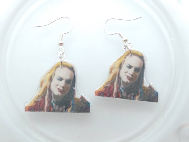 May include: A pair of dangle earrings featuring a printed image of a person with long blonde hair. The earrings are made of clear acrylic and have silver hooks.