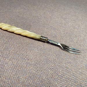 Könnte beinhalten: Kleine Gabel mit dekorativem Griff. Der Griff ist hellcremefarben mit einem gedrehten Design. Die Zinken der Gabel sind silberfarben. Die Gabel ist wahrscheinlich Vintage oder antik.