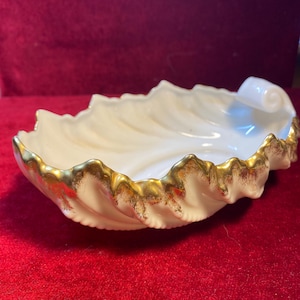 May include: A cream-coloured ceramic dish shaped like a shell with a scalloped edge. The rim of the dish is decorated with a metallic gold trim that follows the curves of the shell, and one end features a small, curled handle detail.