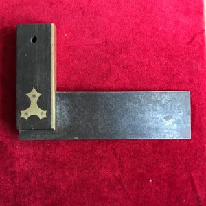 May include: A vintage carpenter's square. The dark wooden handle has brass inlays. The metal blade extends to the right. The square is displayed against a red background, highlighting its design and craftsmanship.